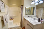 A clean bathroom featuring a walk-in shower with a built-in seat and a beige shower curtain. Next to the shower is a towel rack with folded towels. The vanity has a white countertop with a sink, soap dispenser, and other toiletries. Above the vanity is a large mirror with three light fixtures mounted above it. The walls are painted a light color and there is framed artwork hanging on the wall.
