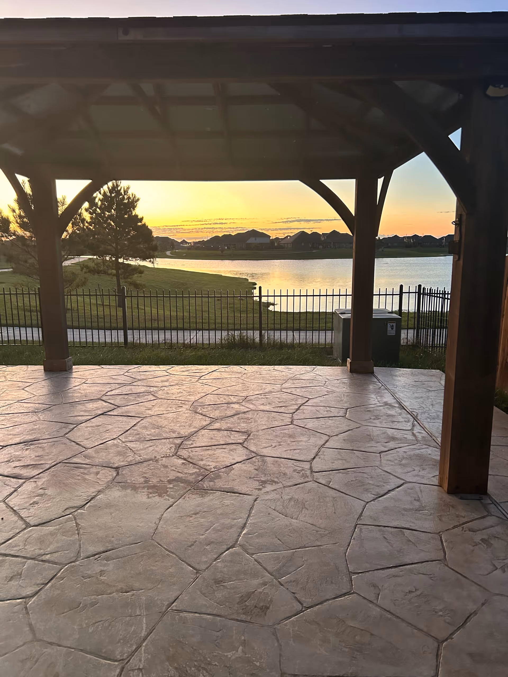 View from under a wooden gazebo with a stone-patterned floor, overlooking a fenced grassy area, a pond, and houses in the distance at sunset.