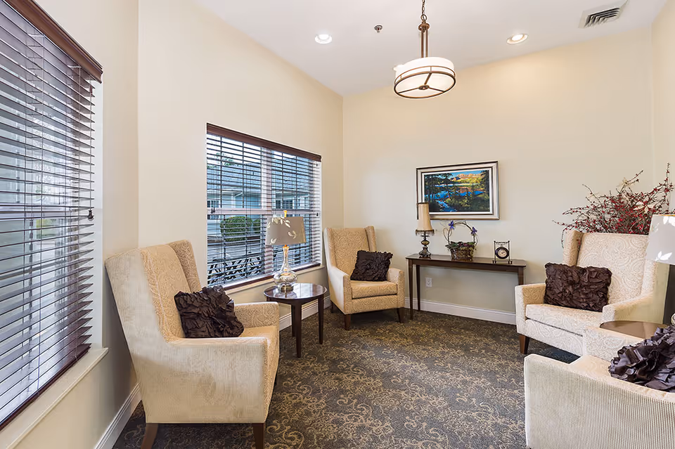 Bright sitting area with upholstered armchairs arranged around side tables, window blinds, a console table with decorative items, and framed wall art.