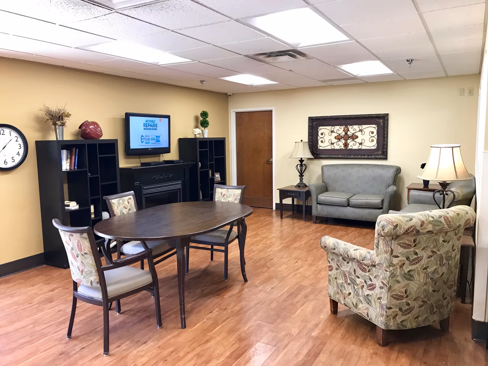 A cozy common area with a wooden oval table surrounded by four chairs with floral upholstery. There are two gray sofas, a floral patterned armchair, two side tables with lamps, a wall clock, two black bookshelves, and a wall-mounted TV displaying a message about activity repairs. The room has wooden flooring and beige walls.