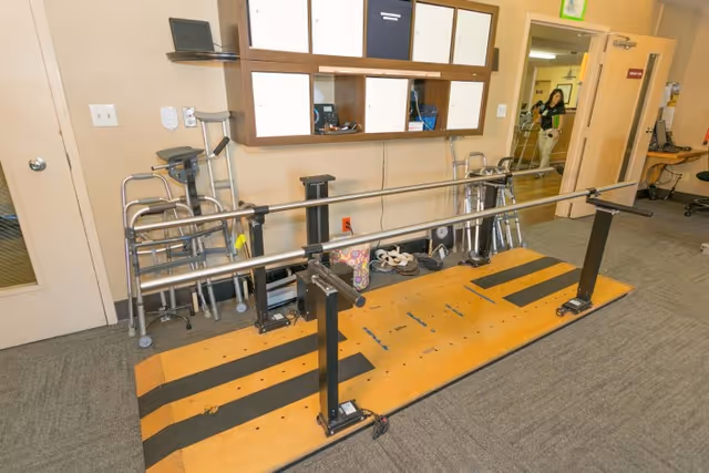 Parallel bars for gait training with walkers and other mobility aids stored against a wall in a rehab area.