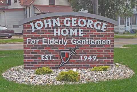Brick sign reading John George Home for Elderly Gentlemen, with crest, on a landscaped lawn.