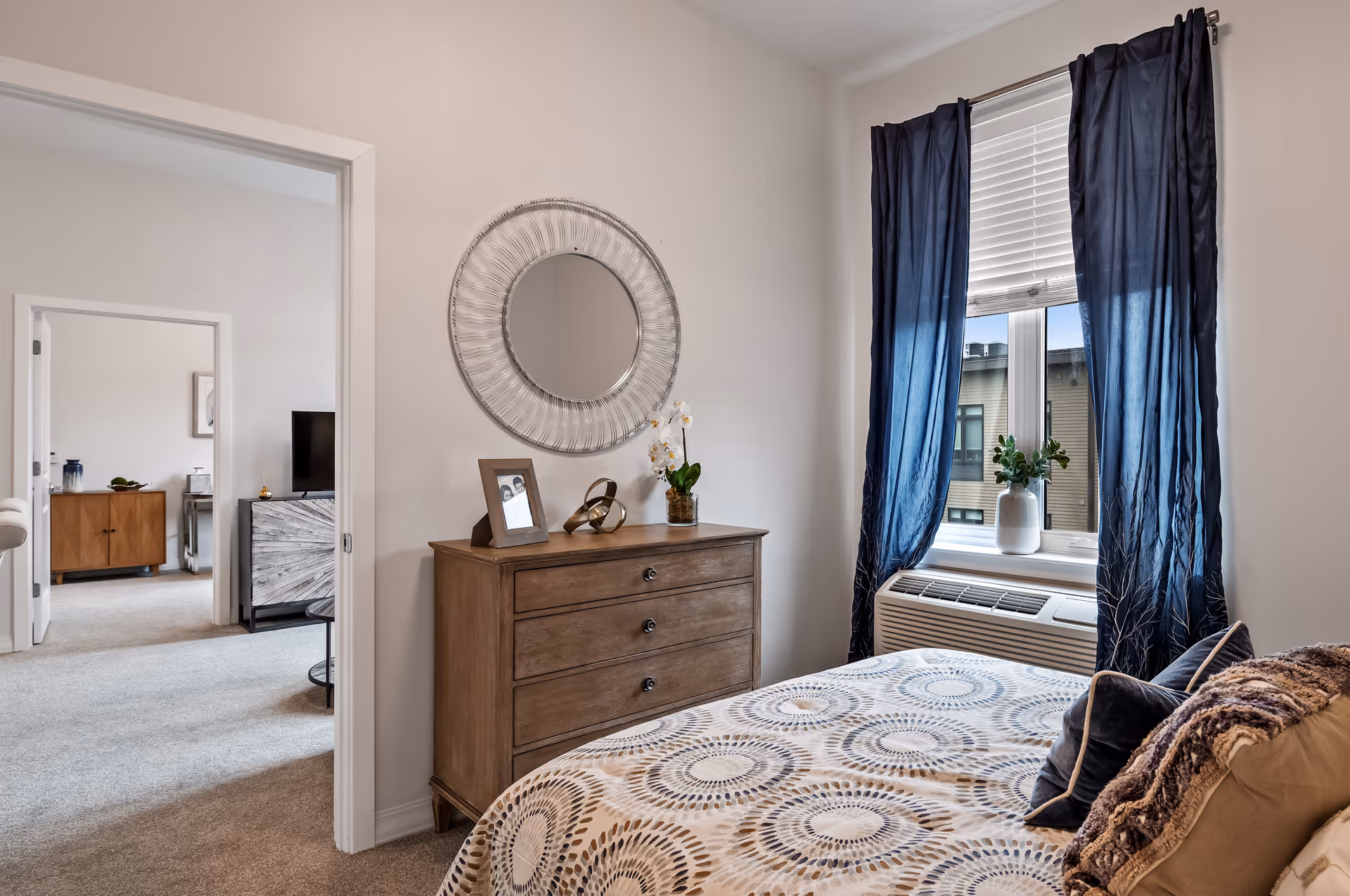 A cozy bedroom with a bed covered in a patterned bedspread and several pillows. Next to the bed is a window with dark blue curtains and a white blind, with a small plant on the windowsill. A wooden dresser with three drawers is against the wall, topped with a round decorative mirror, a framed photo, a small sculpture, and a white orchid plant. The bedroom opens into another room with carpeted floors and furniture visible in the background.