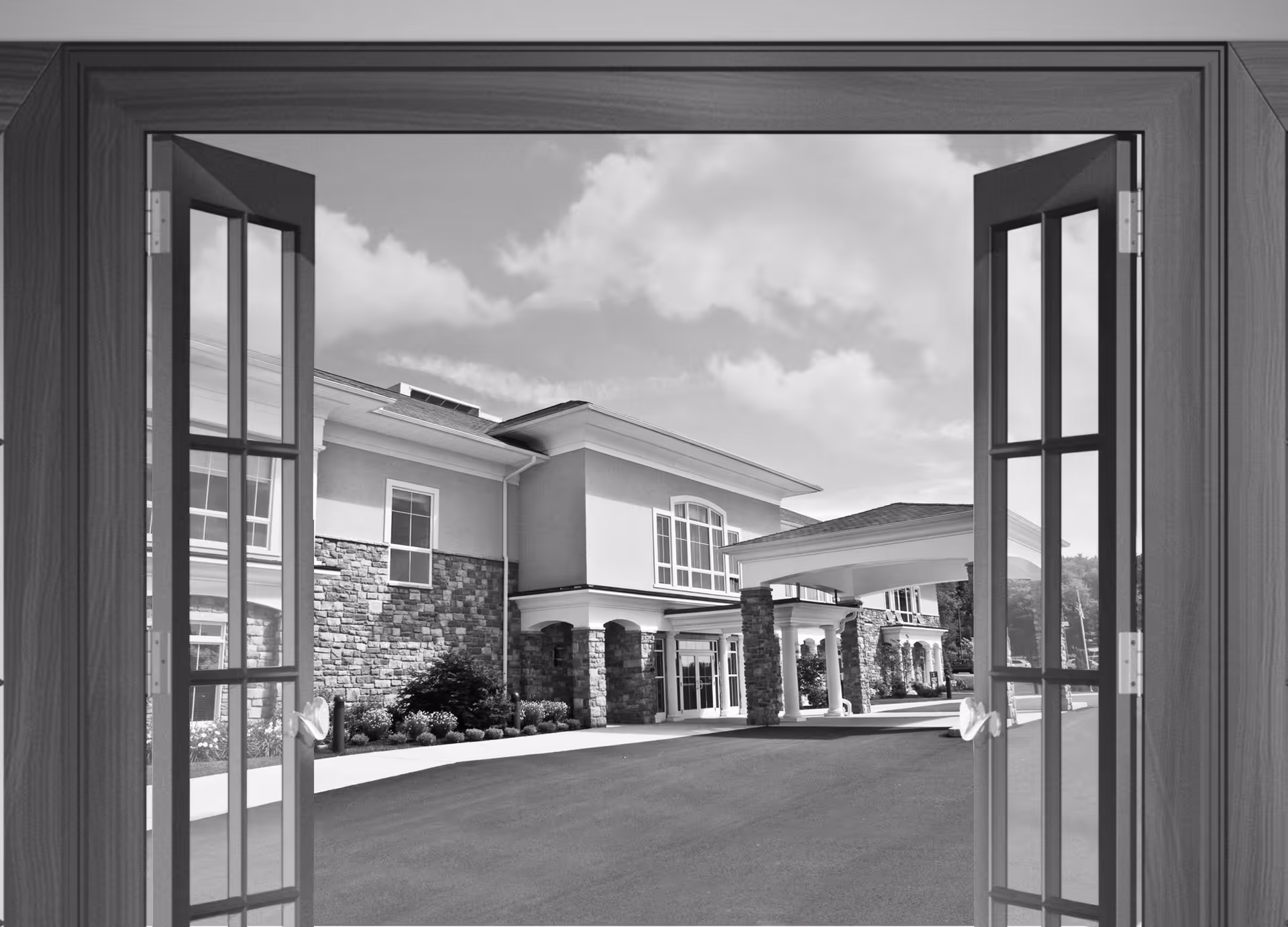 View through open double doors showing the exterior entrance of a senior living facility with stone and stucco walls, large windows, and a covered drop-off area under a partly cloudy sky.