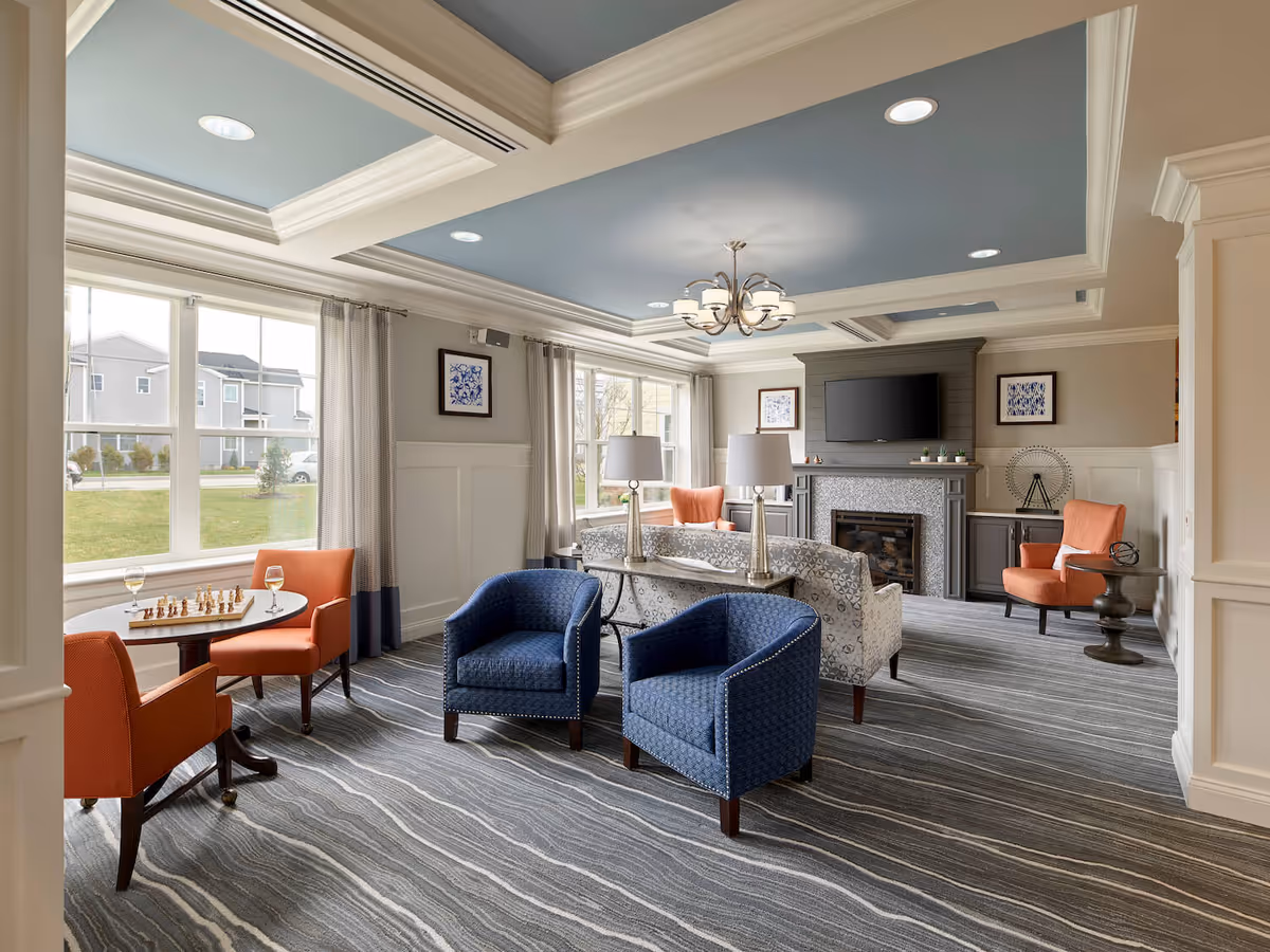 A bright and spacious living room in an assisted living facility featuring large windows with curtains, a patterned carpet, two blue armchairs, two orange armchairs, a small round table with a chessboard and two glasses of white wine, a patterned sofa, two table lamps, a fireplace with a mounted flat-screen TV above it, and framed artwork on the walls.