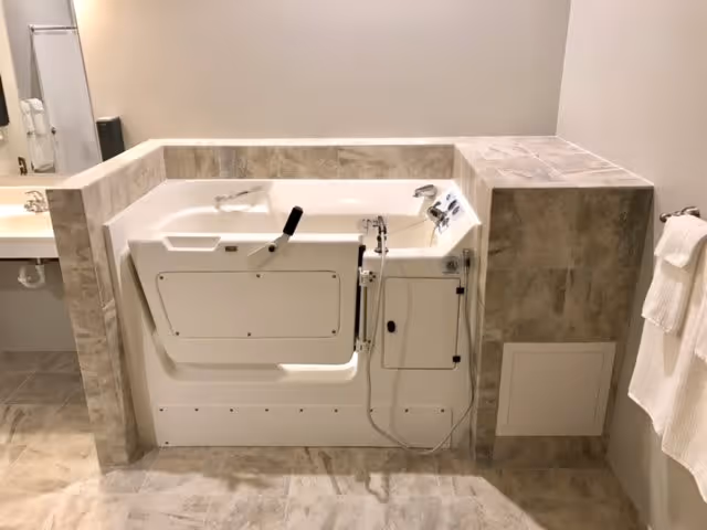 A walk-in bathtub with a side door and handrails, surrounded by beige tiled walls and floor. To the left, there is a sink with a mirror above it, and to the right, a towel rack with white towels hanging.