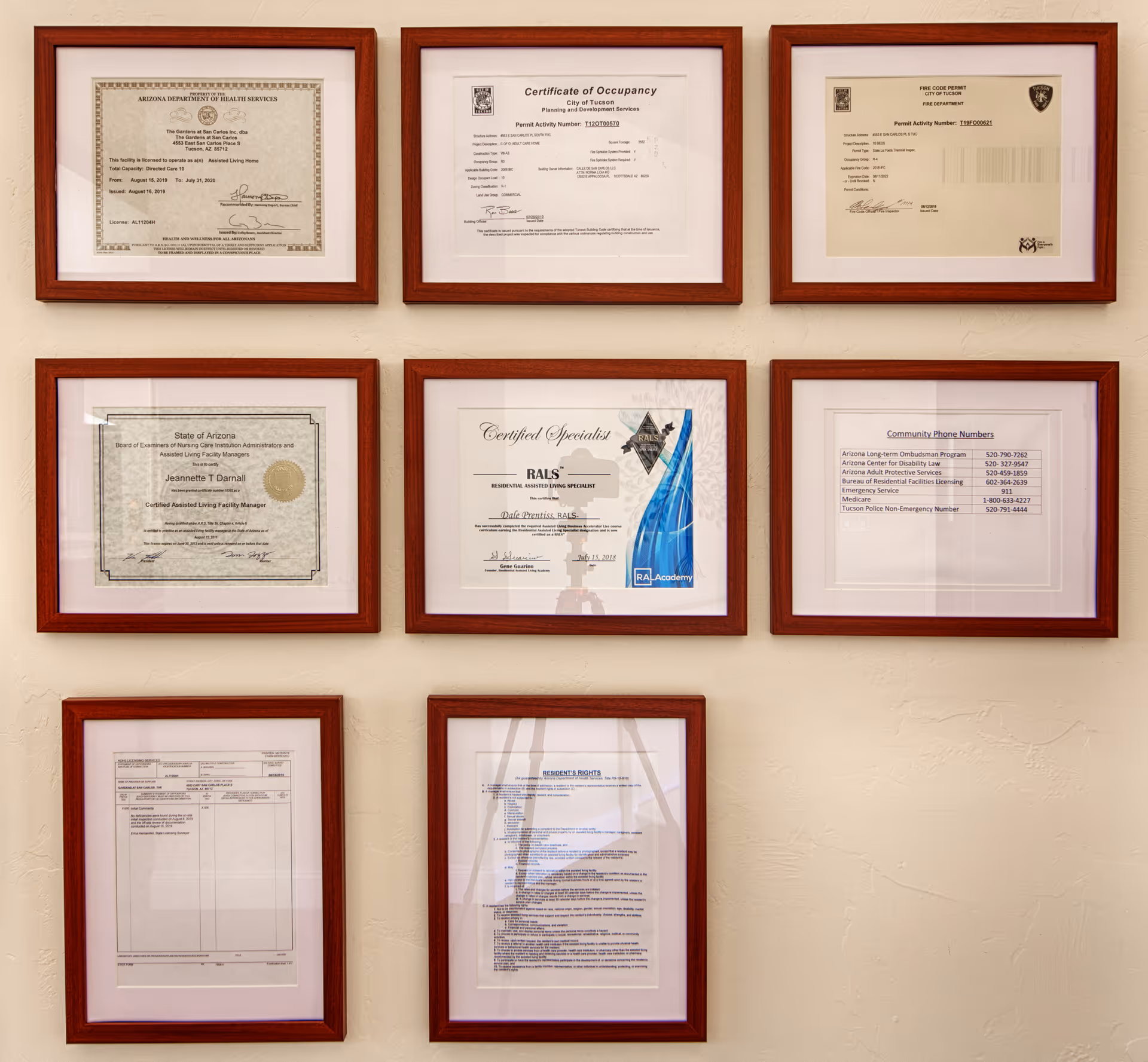 A wall displaying nine framed certificates and documents arranged in a grid. The documents include licenses, permits, certifications, community phone numbers, and residents' rights information, all related to The Gardens at San Carlos assisted living facility.
