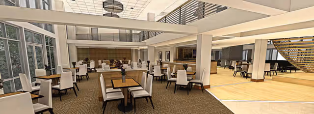 Spacious dining area with multiple wooden tables and white cushioned chairs arranged neatly. Large windows on the left side allow natural light to fill the room. The space features high ceilings, modern light fixtures, and an open layout with a staircase and additional seating area visible in the background.