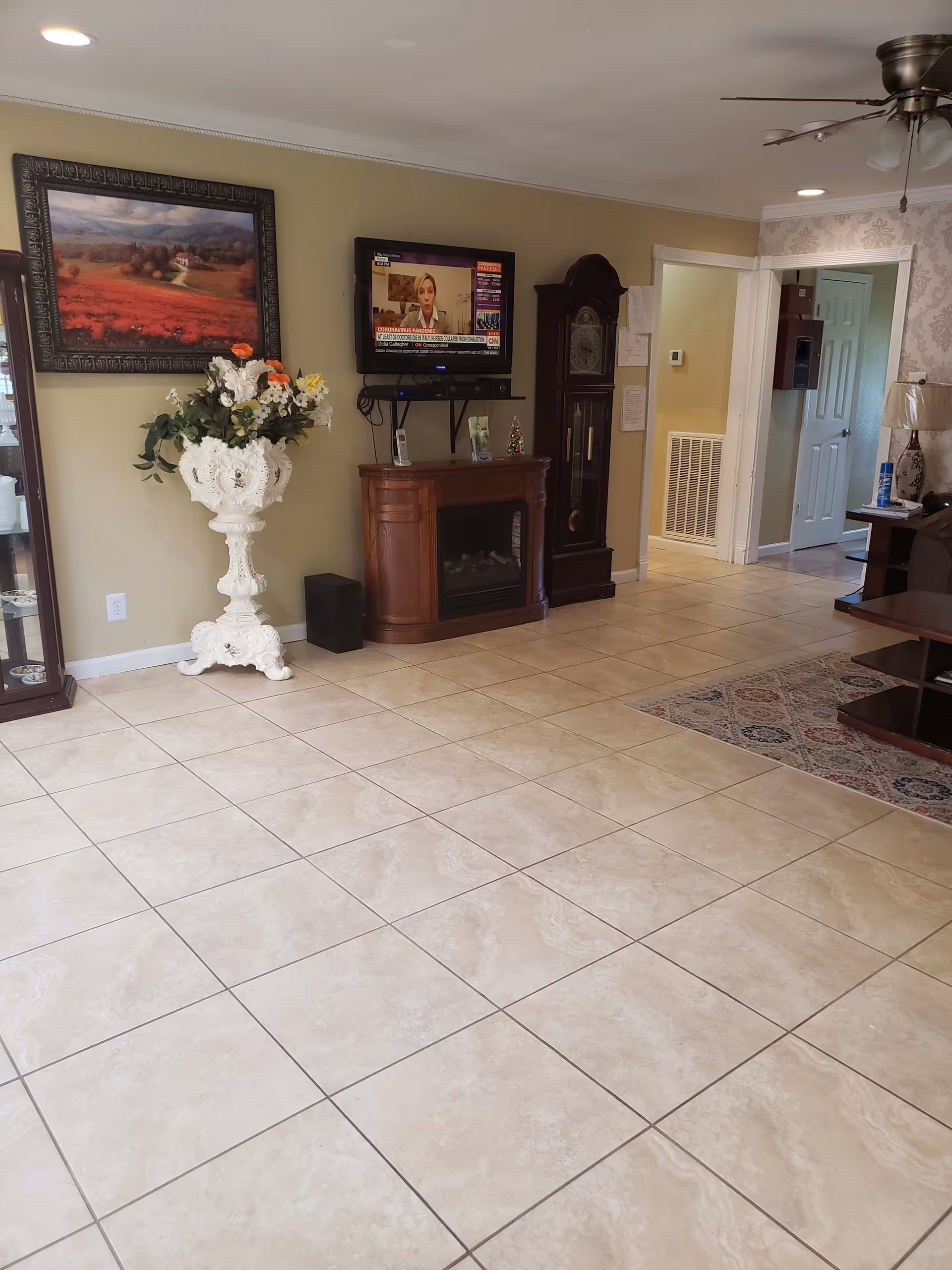 A living room with beige tiled floor, a wall-mounted TV showing a news channel, a wooden grandfather clock, a wooden electric fireplace, a large ornate white vase with flowers, a framed landscape painting, a ceiling fan with lights, and a partial view of a brown sofa and wooden coffee table on a patterned rug.