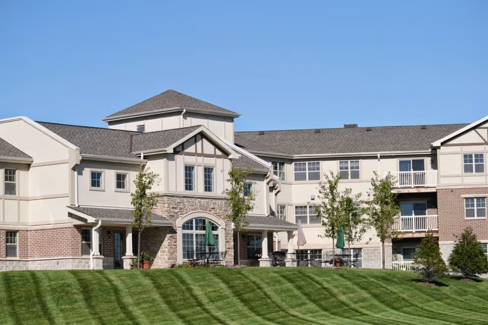 Front exterior of a multi-story senior living building with balconies, patio seating and umbrellas on a manicured lawn under a clear blue sky.