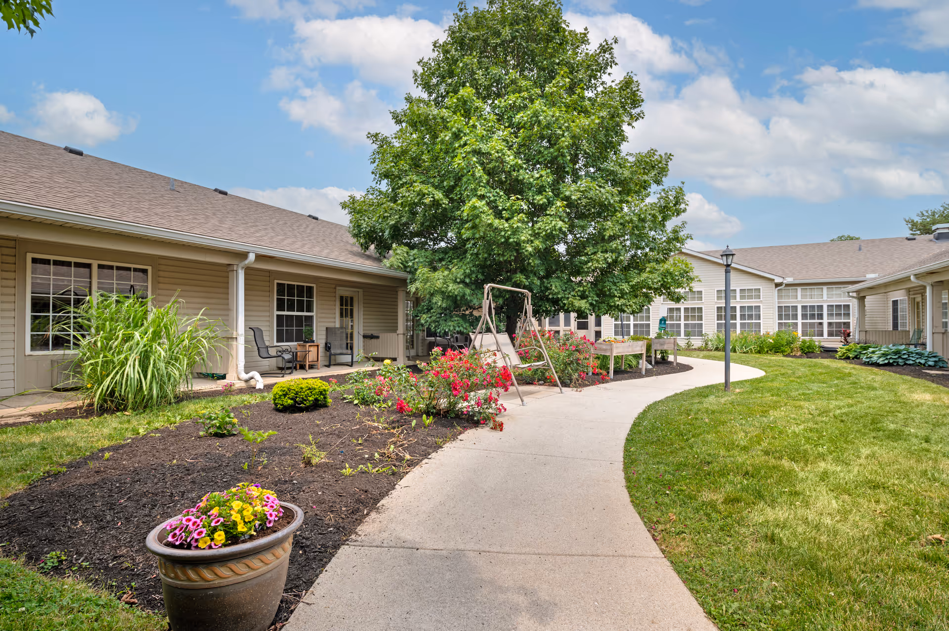 Curved walkway through a landscaped courtyard with flower beds, a bench swing, and surrounding single-story building.