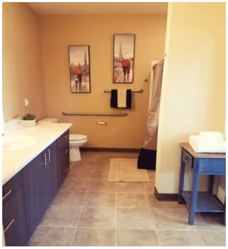 A bathroom with beige walls and tiled floor featuring a long countertop with a sink on the left, a toilet in the center back, two framed pictures above the toilet, a towel rack with two towels, a shower with a curtain on the right, and a small table with folded towels on the far right.