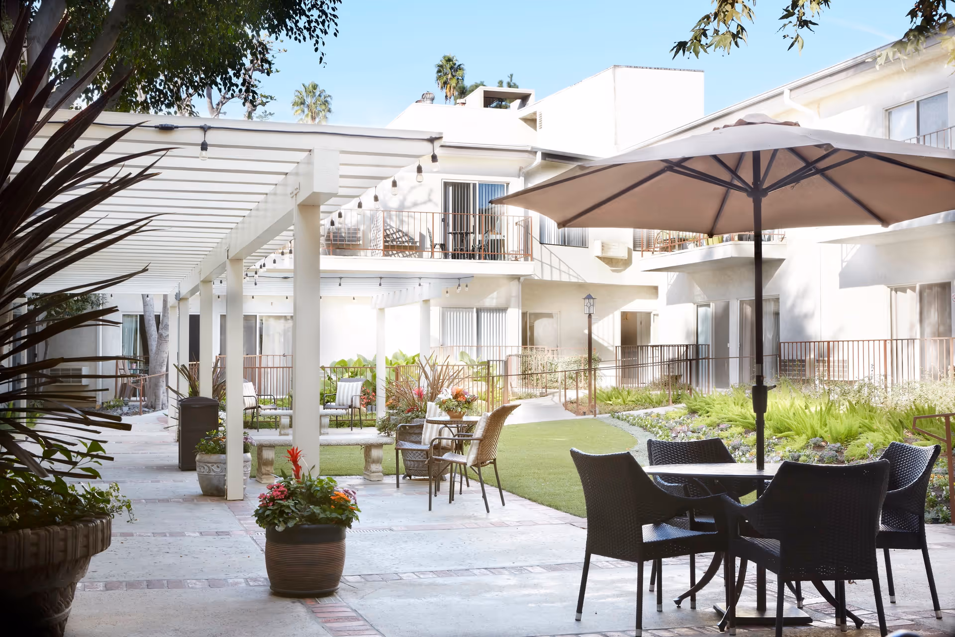 Outdoor courtyard area of a senior living facility with patio tables and chairs, a large umbrella, potted plants, a pergola with string lights, and surrounding two-story building with balconies and windows.