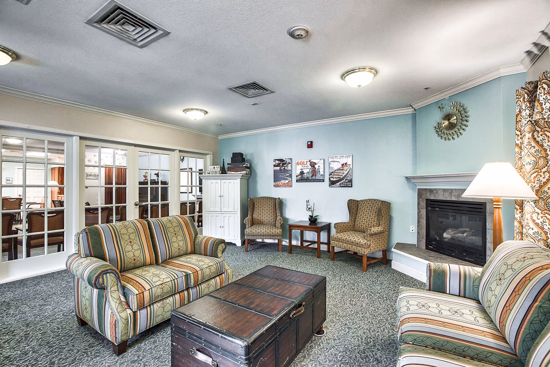 A cozy living room area with two striped upholstered sofas facing a dark wooden trunk used as a coffee table. Two patterned armchairs are placed against a light blue wall with three framed pictures above a small wooden side table. A fireplace with a decorative clock above it is on the right side, next to a floor lamp and patterned curtains. French doors with glass panes lead to another room with a dining table and chairs.