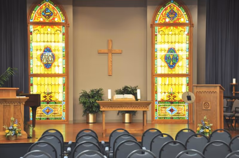 Interior chapel with a wooden altar, cross, stained-glass windows, and rows of chairs.