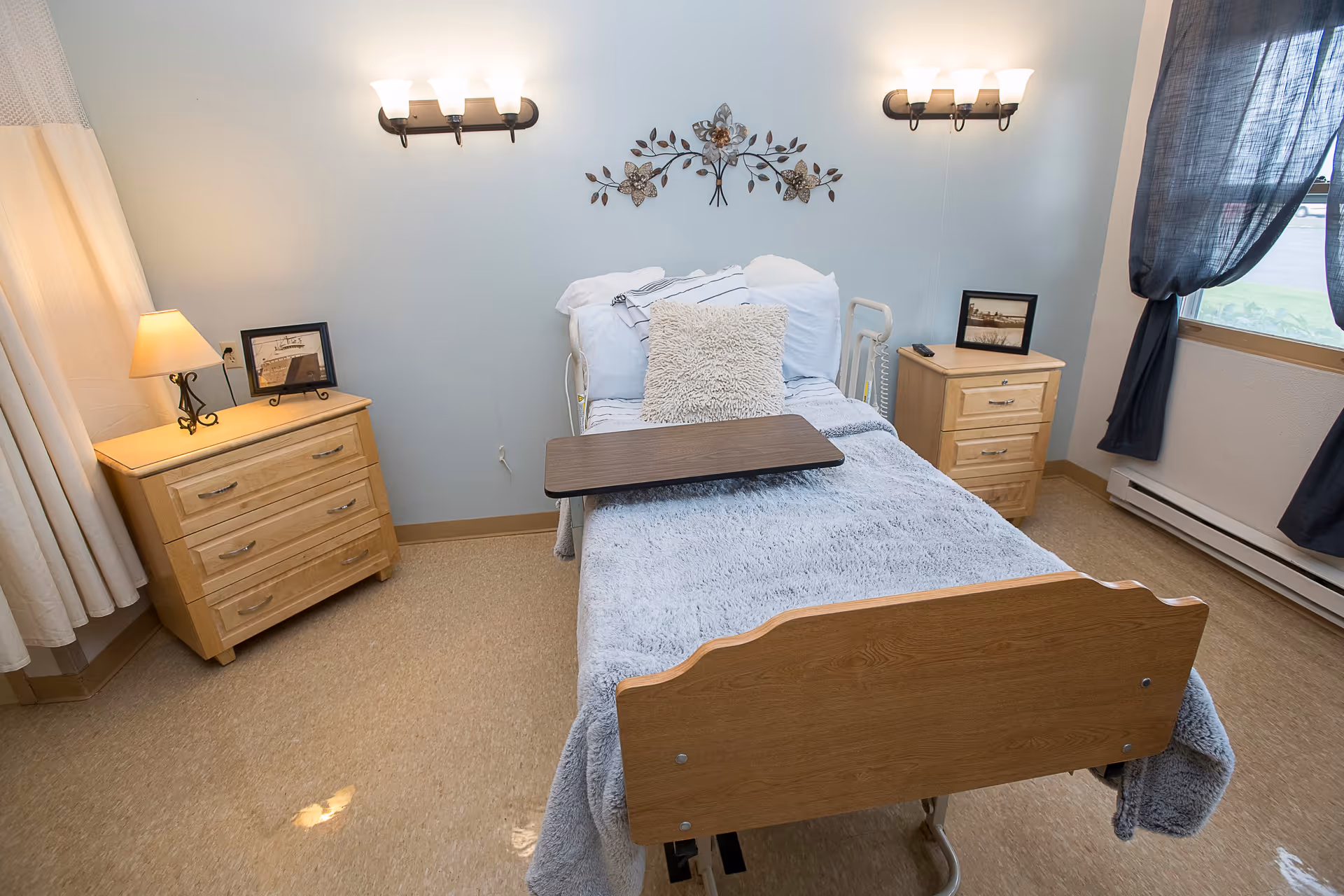 A neatly made single hospital-style bed with a wooden headboard and footboard in a small room. The bed has white and gray bedding with a fluffy pillow and a wooden overbed table. On each side of the bed, there are wooden nightstands with framed pictures and a lamp on the left nightstand. Two wall-mounted light fixtures are above the bed, and a decorative metal wall art piece is centered between them. A window with dark curtains is on the right side of the room, and light-colored curtains are on the left side.