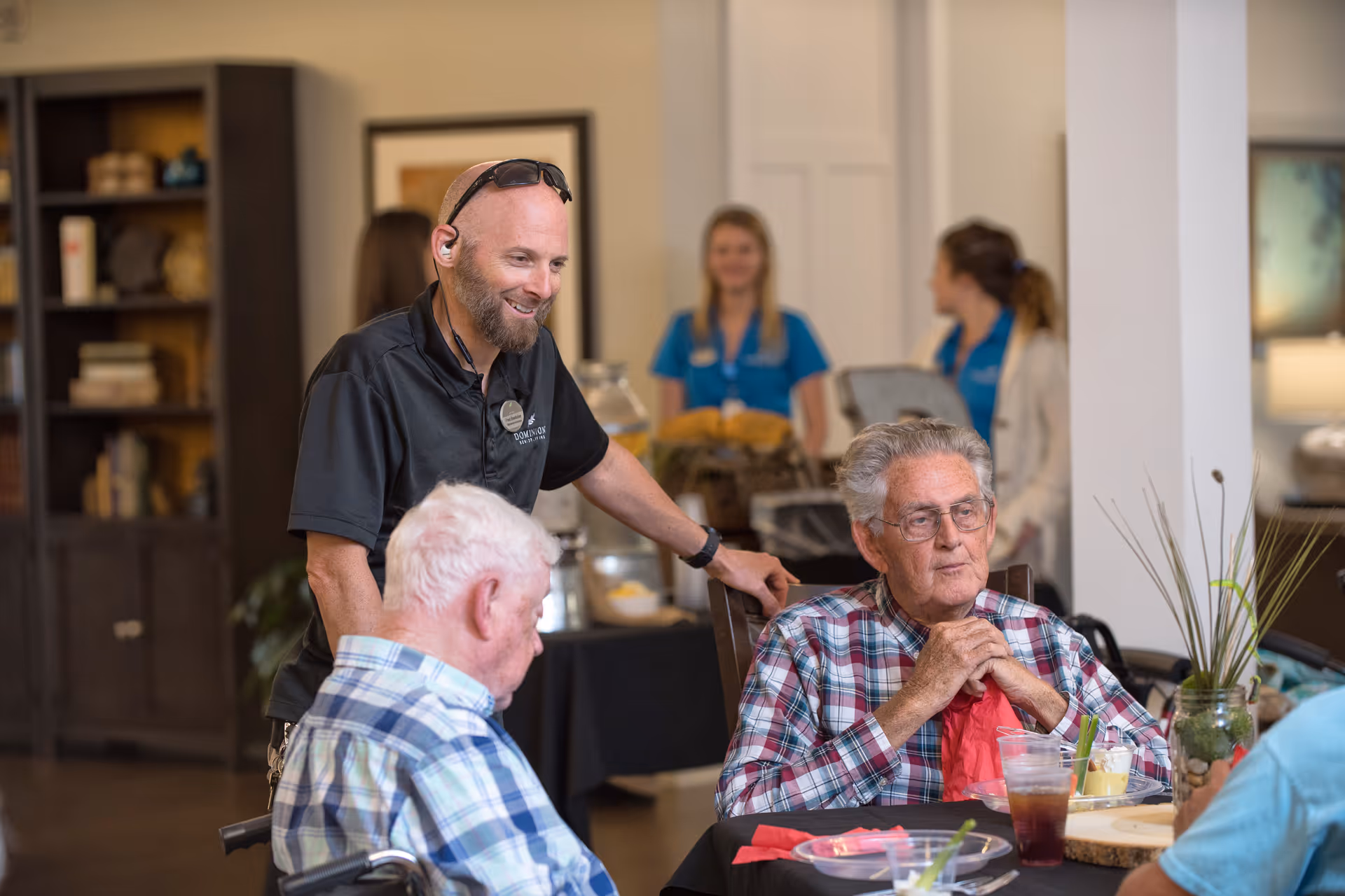 A staff member chats with elderly residents seated at a dining table in a communal area.