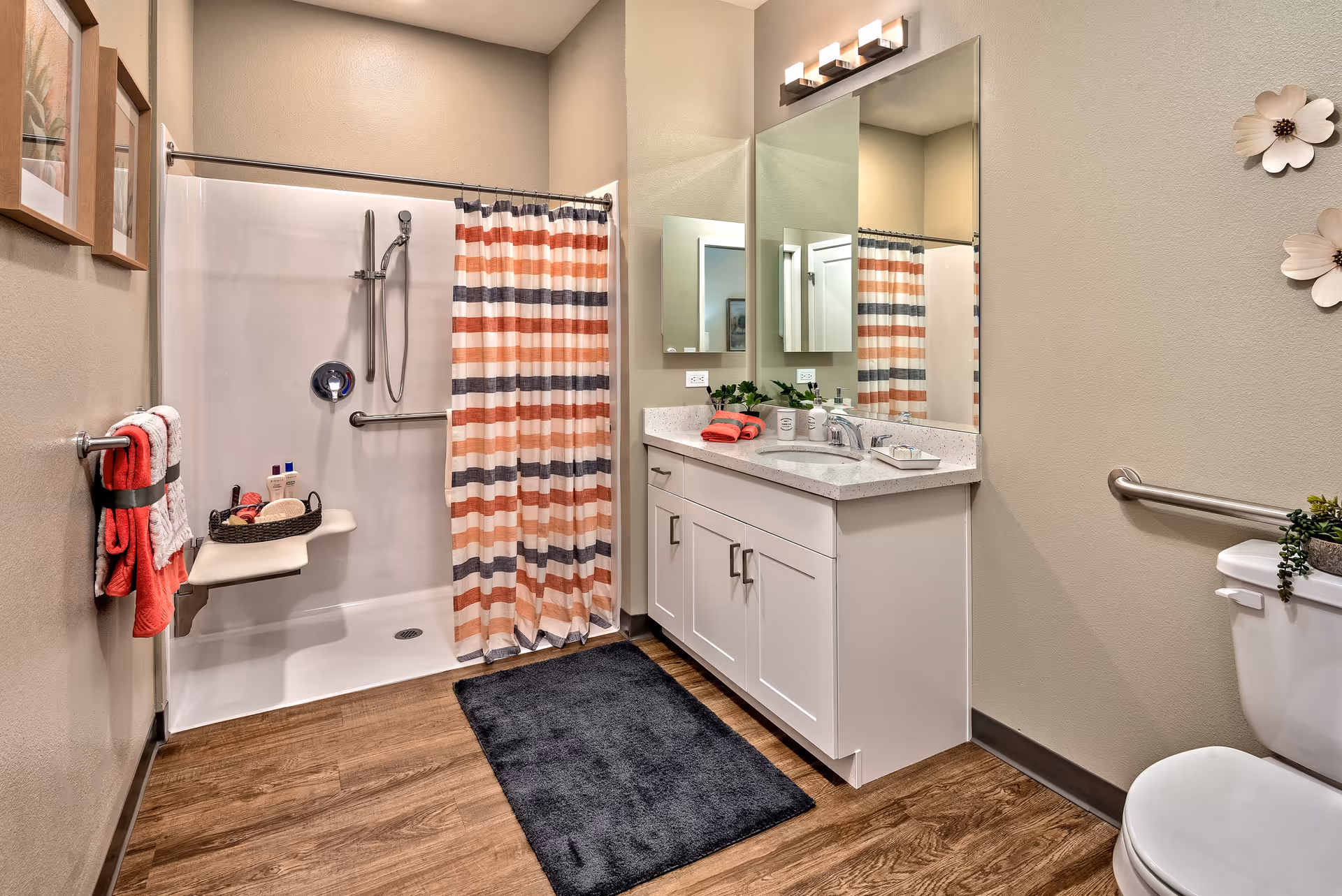 A modern bathroom with a walk-in shower featuring a striped shower curtain in shades of orange, gray, and white. The shower has a handheld showerhead and a fold-down seat. There is a white vanity with a sink, a large mirror, and a light fixture above it. The bathroom also includes a toilet with a small plant on top, a dark gray rug on the wooden floor, and decorative wall art and towels in red and white.