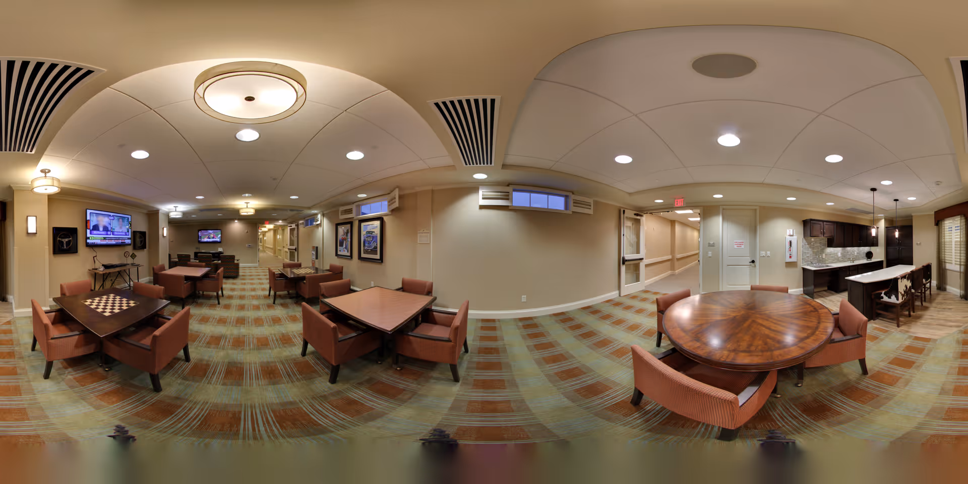 A spacious common area in a senior living facility featuring multiple tables with chairs arranged for socializing and activities. The room has a patterned carpet, beige walls, and a ceiling with recessed lighting and decorative fixtures. There are two wall-mounted televisions, framed artwork, and a kitchen area with a counter and bar stools visible in the background. The space is well-lit and designed for comfort and community engagement.