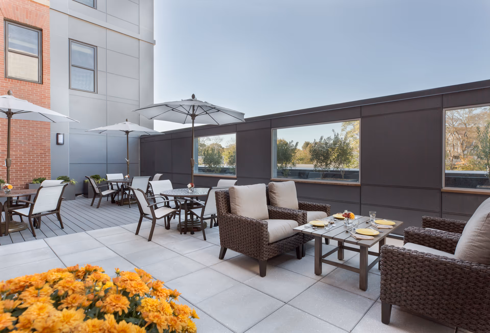 Sunny outdoor patio with wicker seating, dining tables, umbrellas, and potted flowers next to a building.