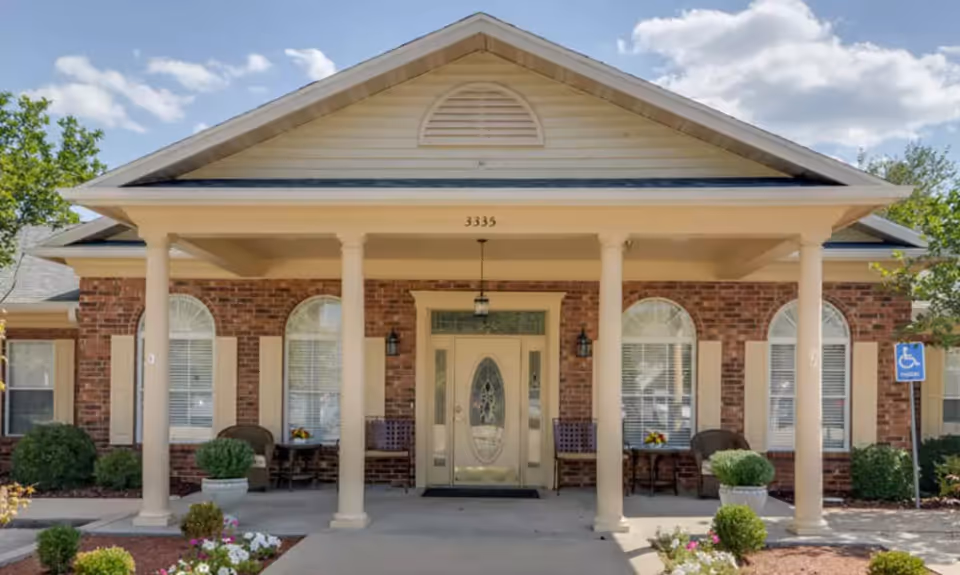 Front exterior view of a single-story brick building with a covered porch supported by four columns. The entrance features a decorative glass door flanked by windows with white shutters. There are chairs and small tables with flower arrangements on the porch. The building number 3335 is displayed above the door. Landscaping includes bushes and flower beds, and a handicap parking sign is visible on the right side.