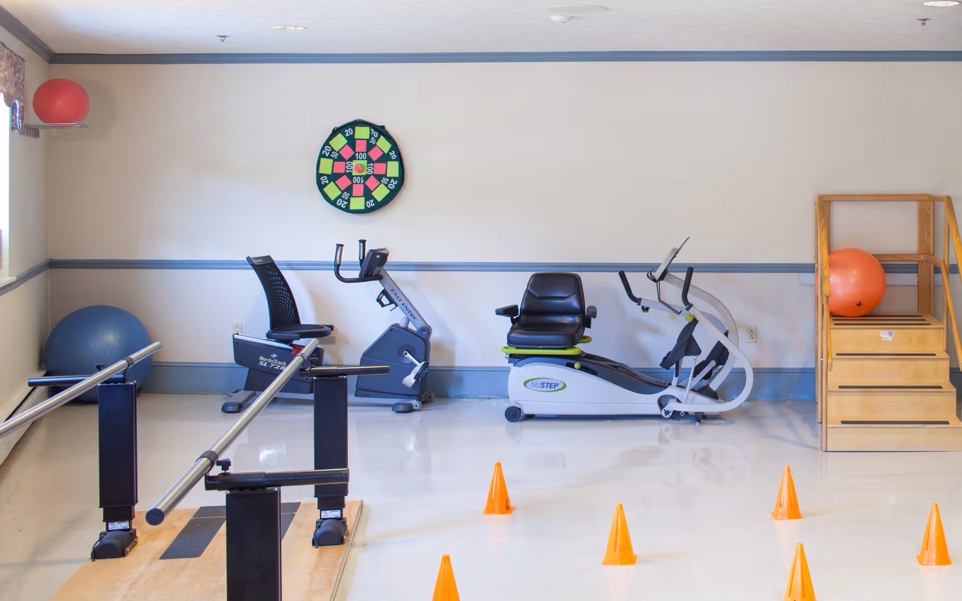 A small exercise room with two stationary recumbent bikes, a set of parallel bars for walking practice, orange cones arranged on the floor, a large blue exercise ball, a red exercise ball on a wooden step platform, and a colorful dartboard on the wall.