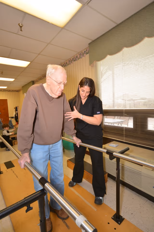 An elderly man wearing a brown sweater and blue jeans is walking with the support of parallel bars in a rehabilitation center. A female healthcare worker in black scrubs is assisting him by holding his arm and providing support. The room has large windows with blinds, fluorescent ceiling lights, and a beige and green color scheme.
