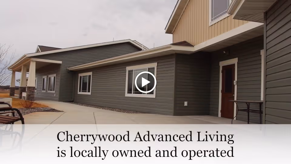 Exterior view of a single-story building with gray siding and white trim, featuring several windows and a door. There is a concrete walkway in front and a bench on the left side. The sky is overcast.