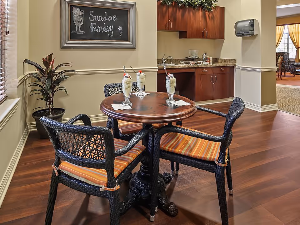 A cozy indoor seating area with a round wooden table and four black wicker chairs with orange striped cushions. On the table are three tall glasses of ice cream sundaes with spoons and cherries on top. Behind the table is a small kitchenette with wooden cabinets, a countertop, and a sink. A chalkboard on the wall reads 'Sundae Funday'. A potted plant is placed near the window with blinds, and a paper towel dispenser is mounted on the wall. In the background, another room with chairs and large windows with yellow curtains is visible.