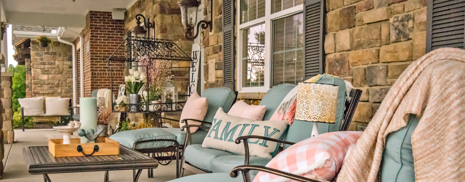 Outdoor patio area with cushioned seating including chairs and a loveseat, decorated with pillows and a throw blanket. There is a coffee table with a wooden tray and decorative items, a lamp, and a stone wall with windows in the background.