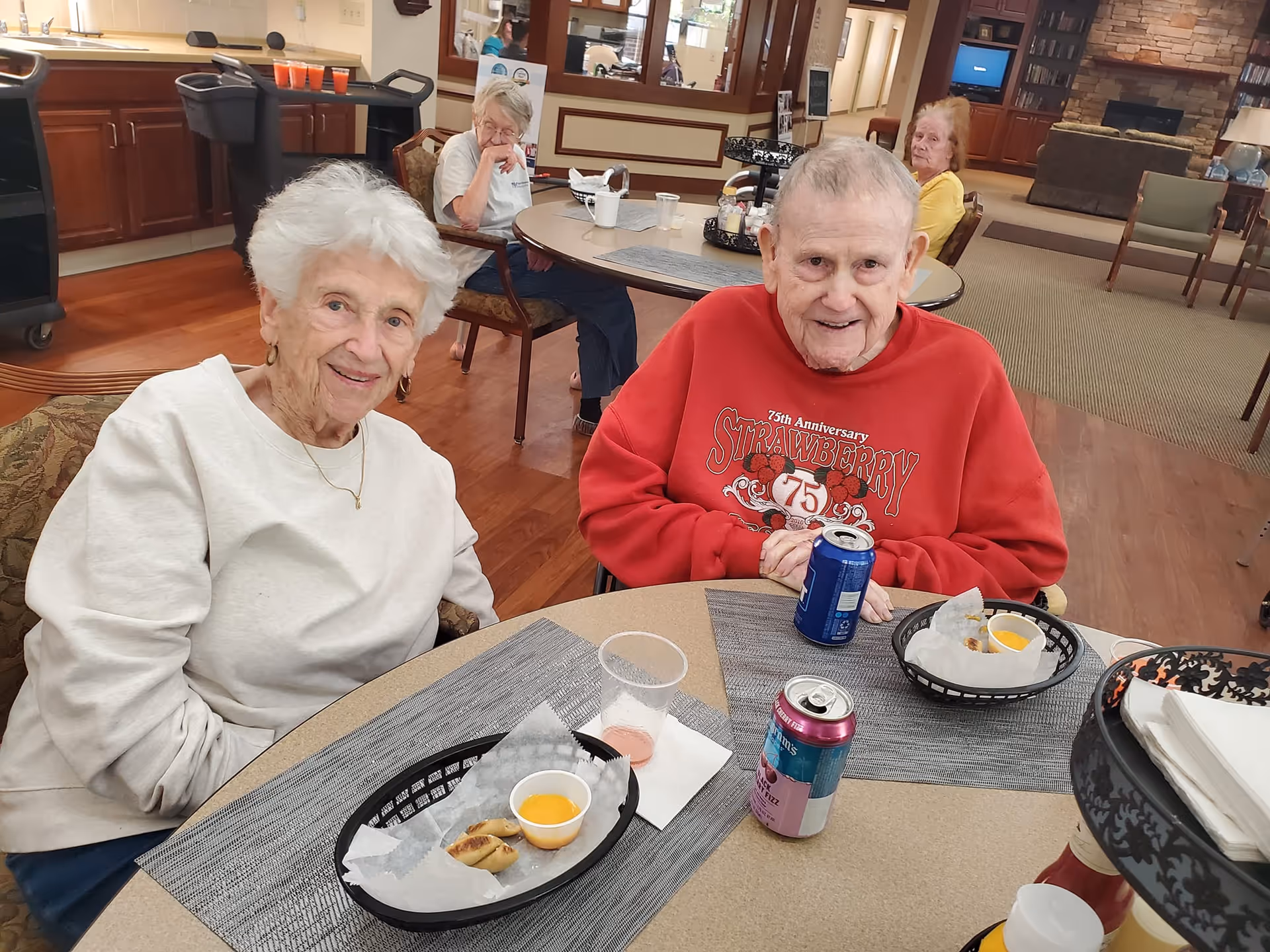 Two elderly individuals sitting at a round table in a communal dining or lounge area. They have trays with small food items and beverages in front of them. In the background, two more elderly individuals are seated at another table. The room has wooden flooring, cabinetry, and comfortable seating, creating a warm and inviting atmosphere.