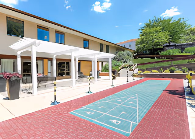 Outdoor patio area at Fairhaven facility with a covered seating area supported by white columns, adjacent to a shuffleboard court with a red brick surface. The building has two stories with multiple windows, and there are trees and greenery on a raised landscaped area in the background under a blue sky with some clouds.