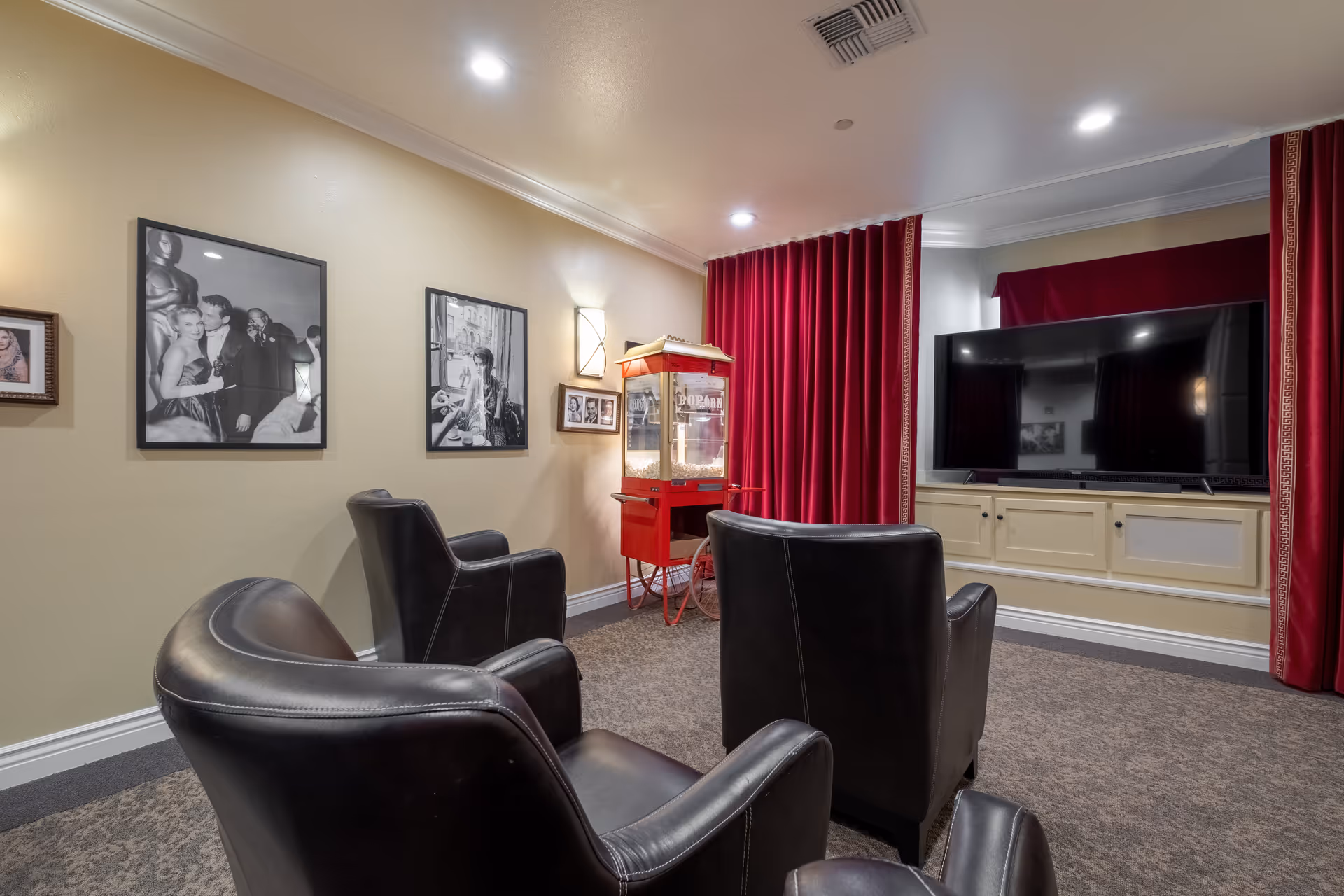 A cozy entertainment room with three black leather armchairs facing a large flat-screen TV mounted on a cream-colored cabinet. The room features red velvet curtains, a vintage-style red popcorn machine, and black and white framed photographs on the beige walls.