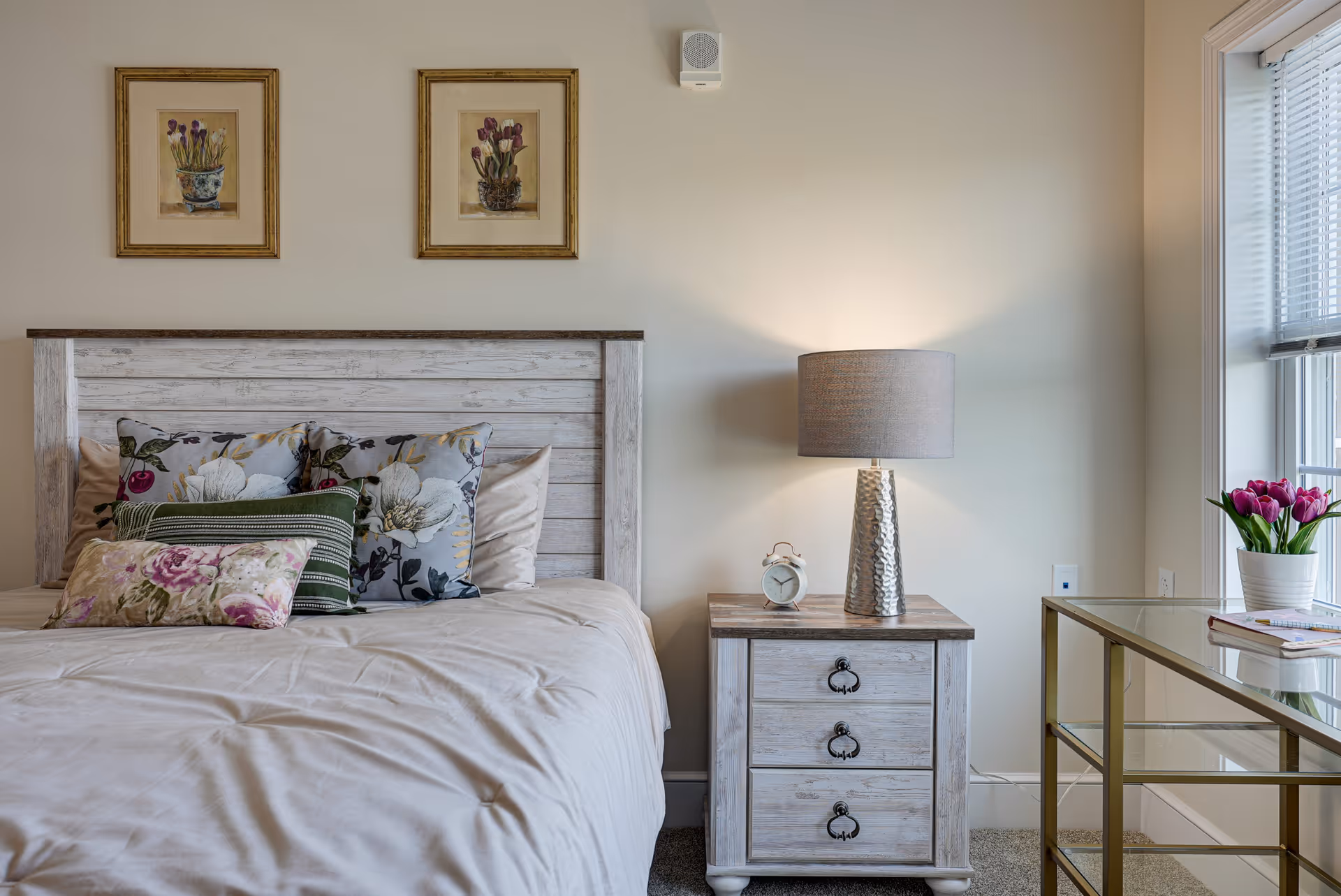 Light, airy bedroom with a whitewashed wooden headboard, floral pillows, a nightstand with lamp and alarm clock, and a glass desk by the window with tulips.