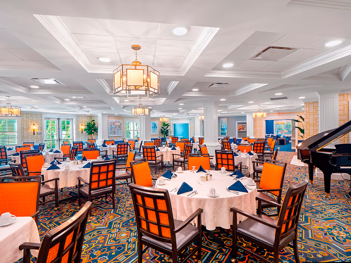 A spacious dining room with multiple round tables covered in white tablecloths, each set with blue folded napkins, cups, and silverware. The chairs have dark wooden frames with bright orange cushions. The room features patterned carpet, white paneled walls with gold accents, large windows letting in natural light, and modern ceiling lights. A black grand piano is visible on the right side of the room.