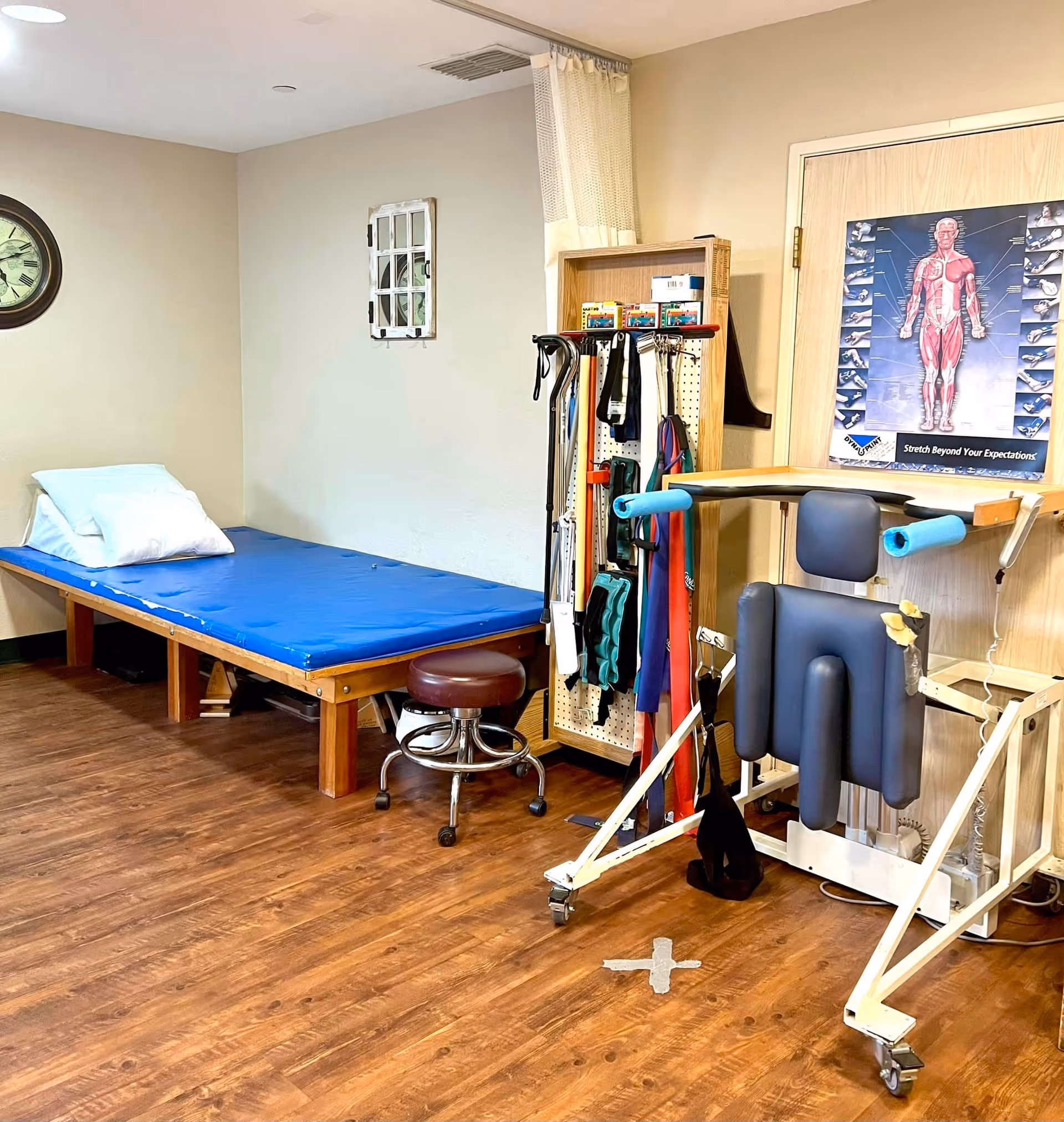A physical therapy room with a blue padded therapy table with pillows, a brown rolling stool, various exercise and therapy equipment including resistance bands and walking canes, and a poster of the human muscular system on the wall.