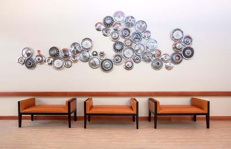 Three orange upholstered benches sit against a wall decorated with a large cluster of metallic hubcap sculptures.