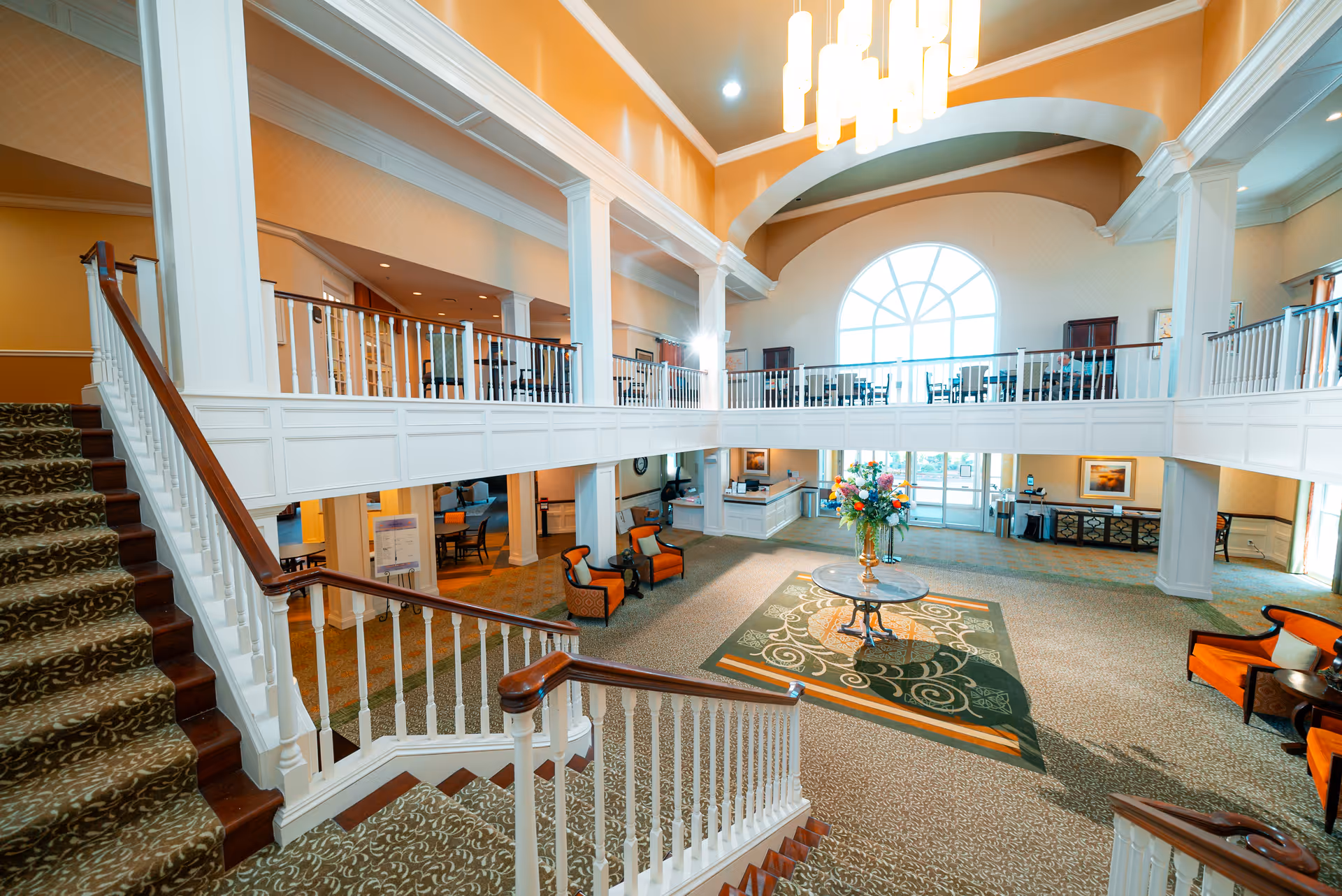 Spacious two-story lobby/atrium with a grand staircase, central table of flowers, seating areas, and a large arched window.