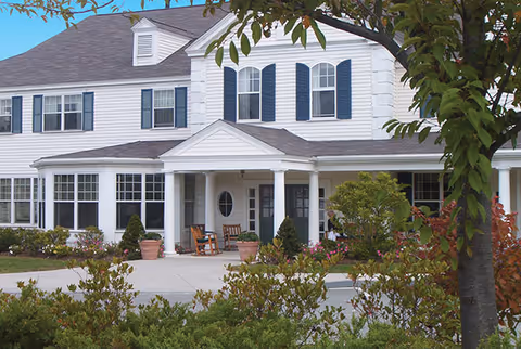 Front exterior view of a two-story white building with blue shutters, a covered porch with rocking chairs, potted plants, and landscaped greenery in the foreground.
