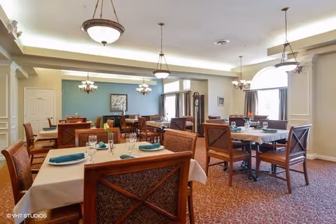 A spacious dining room with several tables covered in beige tablecloths, each set with glassware and teal napkins. The room features warm lighting from hanging chandeliers, patterned carpet, and large windows with curtains allowing natural light to enter. The walls are painted in soft neutral tones with a teal accent wall in the background, decorated with framed artwork and a grandfather clock.