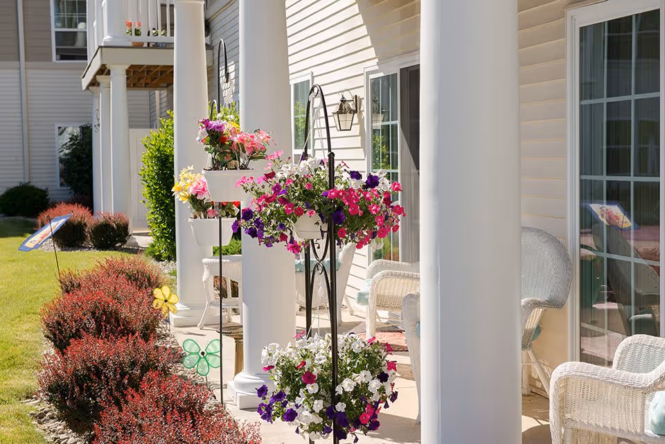 A sunny outdoor patio area with white columns and beige siding. There are hanging flower baskets with colorful pink, purple, and white flowers, white wicker chairs with light blue cushions, and neatly trimmed red bushes along the edge of the patio. Sliding glass doors lead into the building.