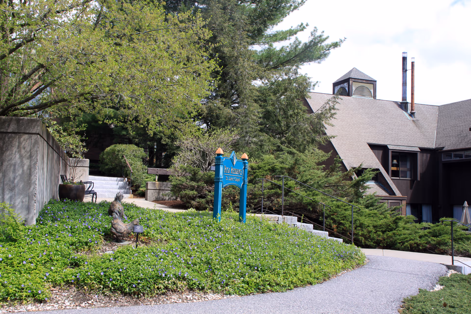 Front exterior of a senior living facility with landscaped greenery, a paved walkway, stairs, and a blue sign in front of a brown building.