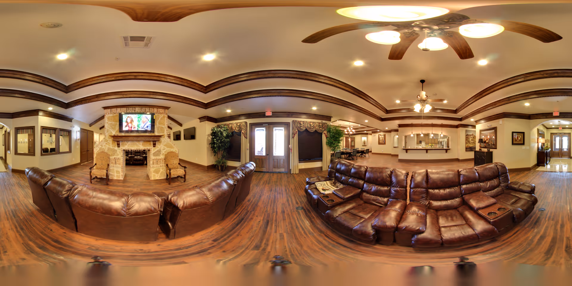A spacious and warmly lit common area in Avalon Memory Care featuring two large brown leather sectional sofas facing each other on a wooden floor. The room has a stone fireplace with a TV mounted above it, two armchairs beside the fireplace, ceiling fans with lights, and decorative crown molding. In the background, there is a reception or service counter area with pendant lights, framed artwork on the walls, and large windows with curtains.
