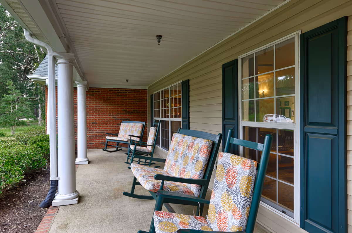 Covered porch area with several green rocking chairs featuring colorful floral cushions, white columns supporting the roof, beige siding with green window shutters, and a brick wall at the far end. Green bushes and trees are visible on the left side.