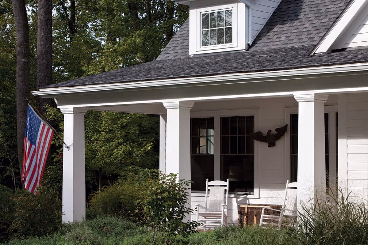 Front porch of a white house with two white rocking chairs, a wooden chest, an American flag mounted on a pole, and green bushes and trees surrounding the porch.