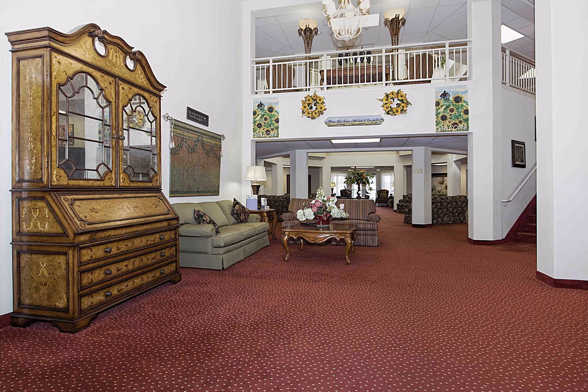 A spacious and well-lit living room area in a senior living facility with red patterned carpet, antique wooden cabinet, green and striped sofas, a wooden coffee table with a floral arrangement, and decorative wall hangings including sunflower wreaths and welcome signs. There is a second-floor balcony with white railings overlooking the room.