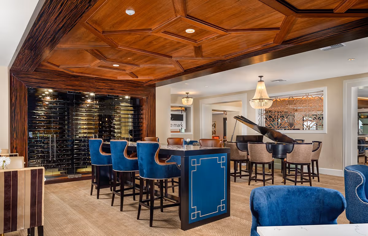 Elegant interior lounge area with a wooden coffered ceiling, a glass-enclosed wine display, blue and brown high-back chairs around a tall table, a grand piano with beige chairs, and chandeliers providing warm lighting.