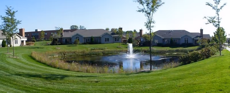 A scenic outdoor view of Glacier Hills Senior Living Community featuring a small pond with a water fountain in the center, surrounded by well-maintained green lawns and several single-story residential buildings in the background under a clear sky.