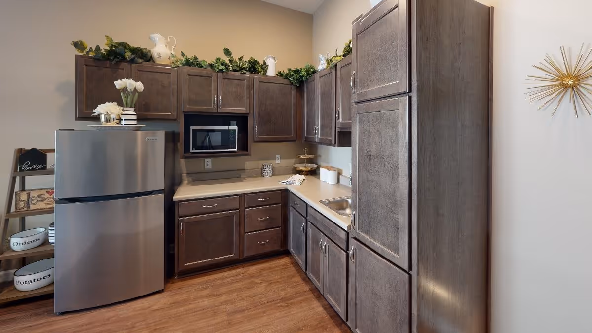 A modern kitchen with dark wood cabinets, a stainless steel refrigerator, a microwave built into the cabinetry, a small sink, and a countertop with decorative items. There is a wooden shelf on the left side holding bowls labeled 'Onions' and 'Potatoes' and other decorative pieces. The floor is wood, and the walls are painted beige with a gold starburst wall decoration on the right.