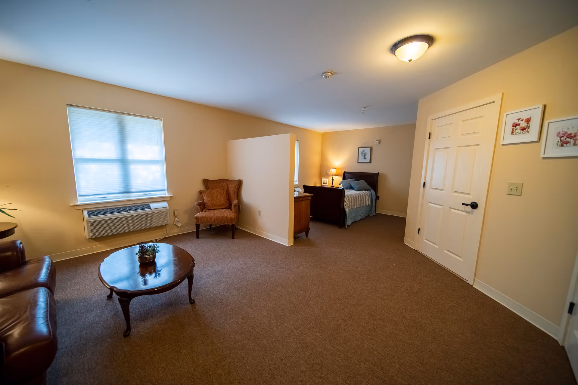A senior living facility room with a small sitting area and a sleeping area separated by a partial wall. The sitting area has a leather sofa, a round wooden coffee table with a small plant, and an upholstered armchair near a window with closed blinds and an air conditioning unit below it. The sleeping area contains a single bed with a wooden headboard, a nightstand with a lamp, and framed artwork on the walls. The room has beige walls, carpeted floor, and a ceiling light fixture.